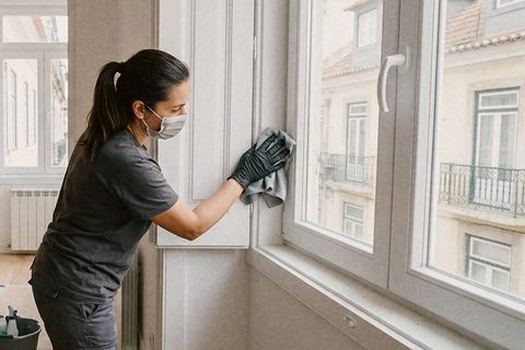 Profissional de máscara e luvas a limpar o pó de obra no aro de uma janela nova, num apartamento acabado de remodelar